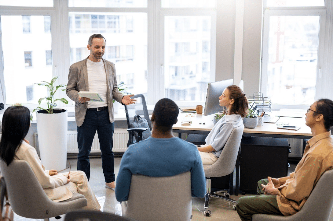 A man stands and speaks to four seated colleagues in a modern office with large windows, desks, and plants. The group appears to be having a discussion or meeting, with everyone focused on the speaker.