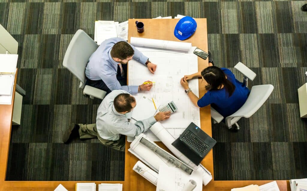 Three people sit around a table covered with blueprints, papers, a laptop, and a blue hard hat, collaborating on a project in an office with a striped carpet.