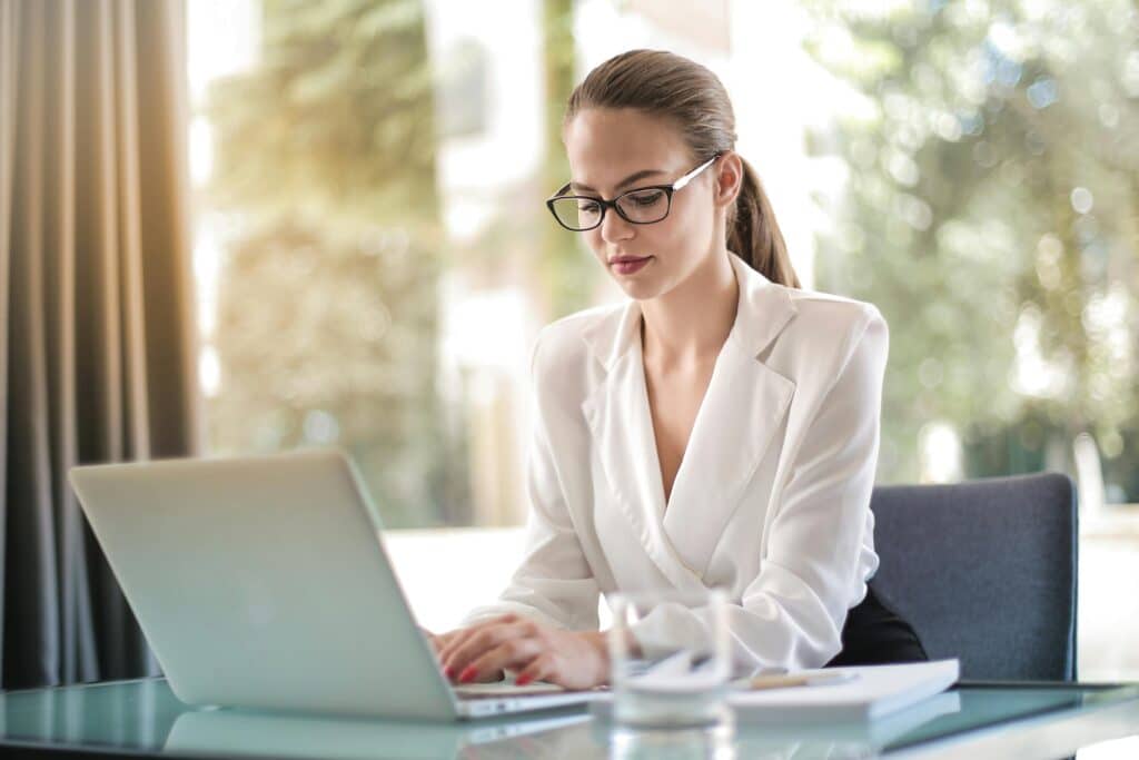 A woman wearing glasses and a white blazer is sitting at a glass table, typing on a laptop. She is focused on her work, with documents and a glass of water beside her, and greenery visible through the window in the background.