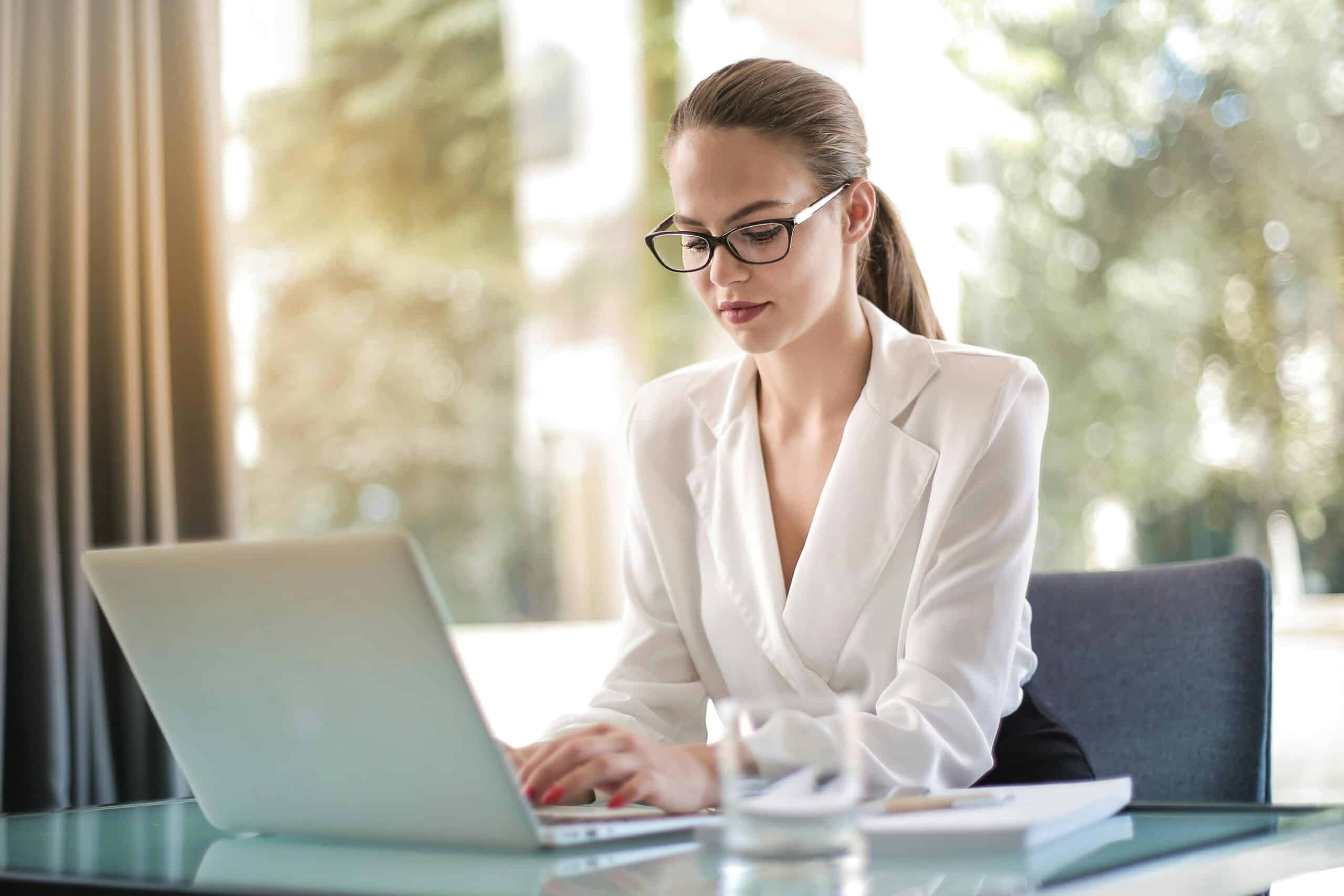 A woman wearing glasses and a white blazer is sitting at a glass table, typing on a laptop. She is focused on her work, with documents and a glass of water beside her, and greenery visible through the window in the background.