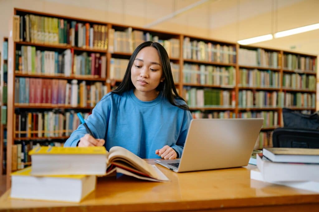 A young woman in a blue sweater studies at a library table, writing in a notebook with a laptop open in front of her and several books stacked nearby, bookshelves in the background.