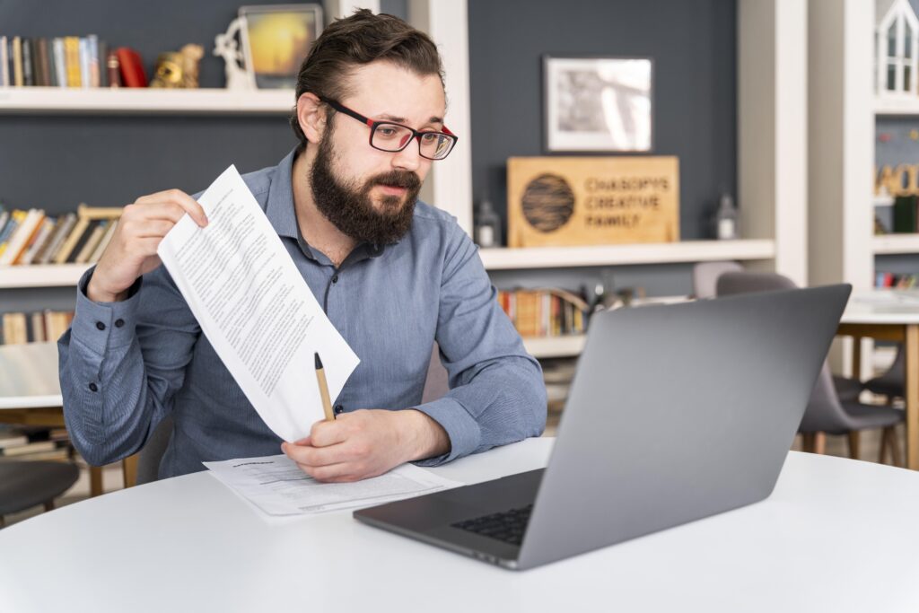 A man with glasses and a beard sits at a table holding up a document and a pen while looking at a laptop, appearing to explain something, with bookshelves in the background.