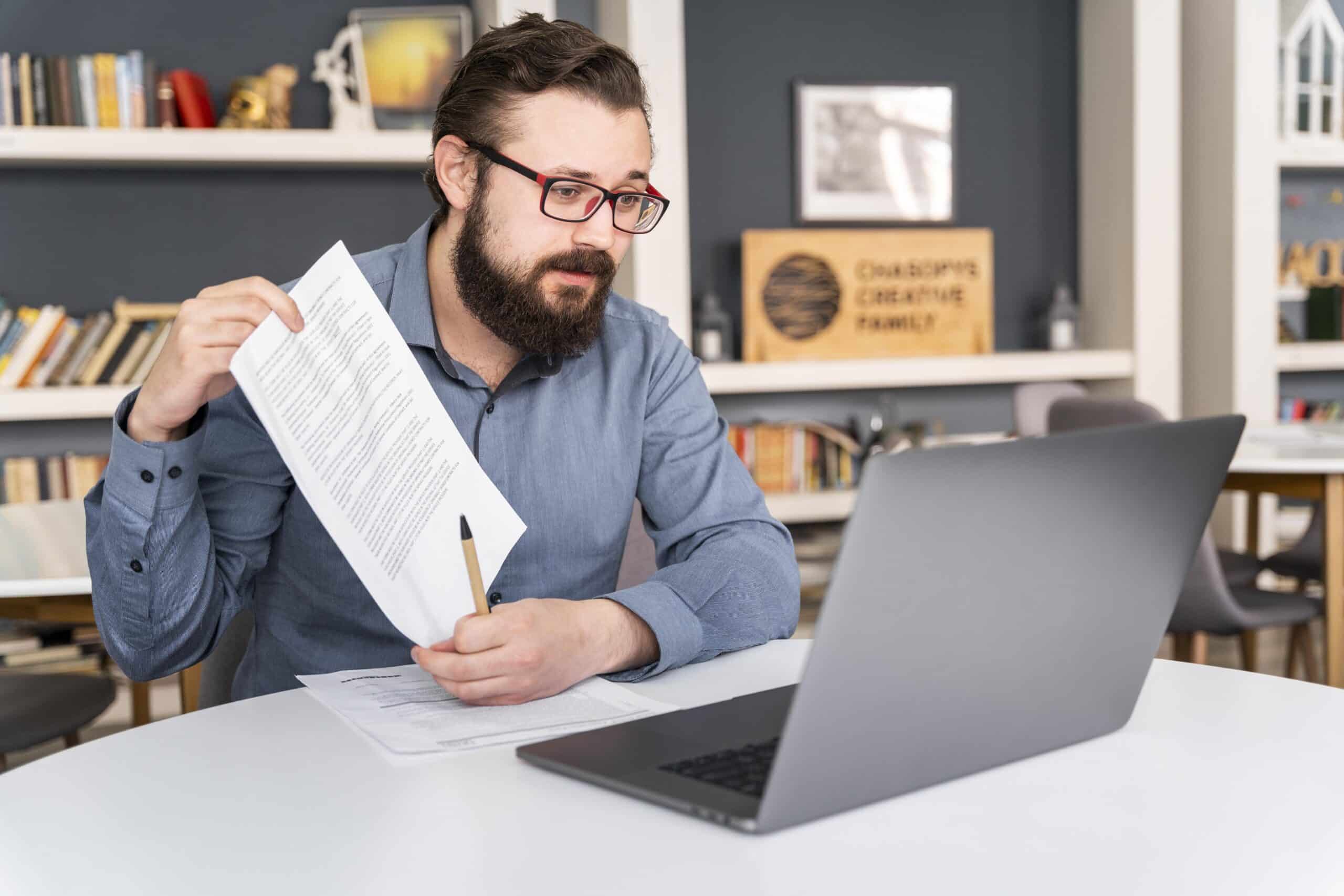 A man with glasses and a beard sits at a table holding up a document and a pen while looking at a laptop, appearing to explain something, with bookshelves in the background.