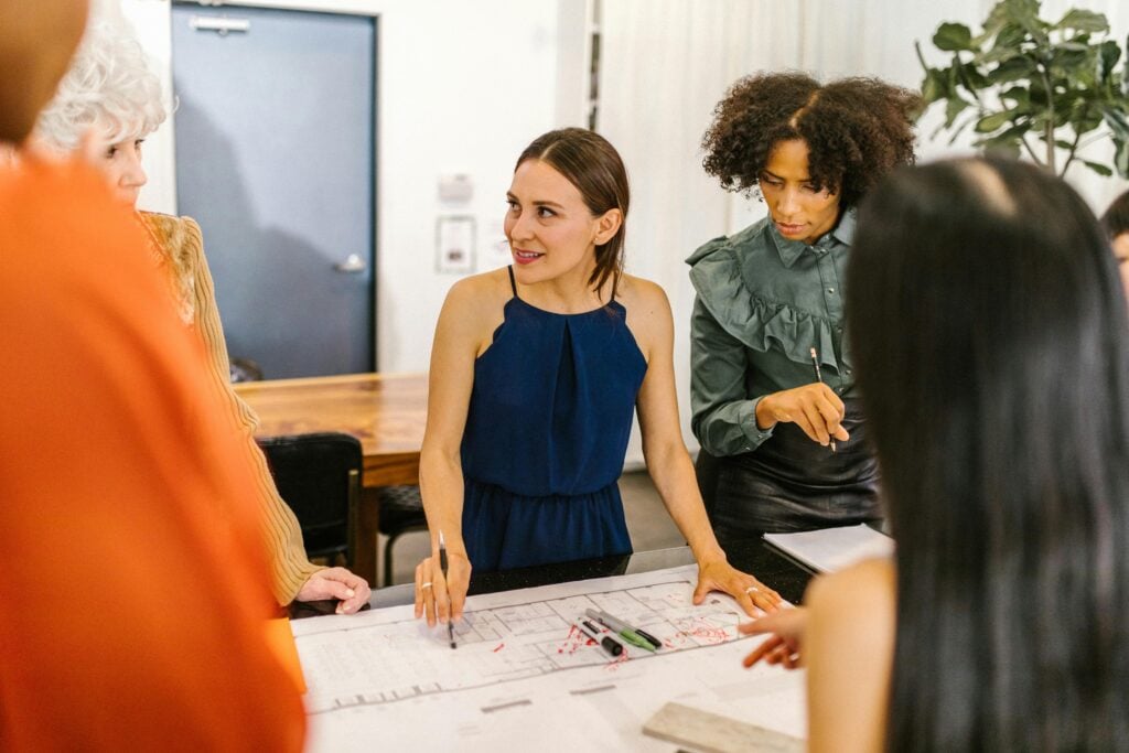 A group of four people stand around a table covered with architectural plans and markers, discussing a project in a modern office setting. One woman in a blue dress is speaking and gesturing with a pen.